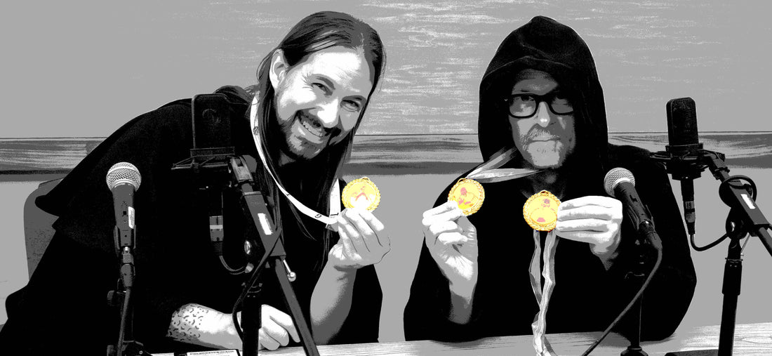 Two men in robes holding up medals sitting behind microphones at a desk, the man on the left is smiling and leaning in while the man on the right is stone faced and overly serious.